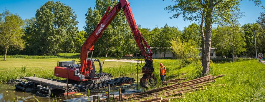 Rode kraan werkt vanaf het water aan versterking van de kade. Hij verplaatst grote houten palen. Er staat een werkman aan de oever.