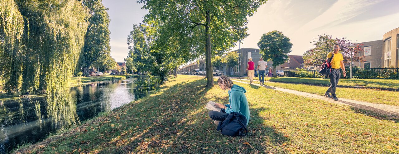 Jonge vrouw met koptelefoon leest een boek aan de waterkant, op het gras. Het is herfst, de bladeren vallen al van de bomen. Op het voetpad lopen twee oudere mensen met nordic walking-stokken en er loopt een man voorbij met een sporttas. De zon schijnt